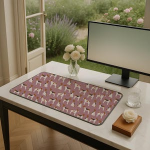May include: A rectangular desk mat featuring a repeating pattern of dogs on a mauve background. The mat is on a white desk, with a computer monitor, a vase of white roses, and a glass of water. The mat is about 76 cm long.