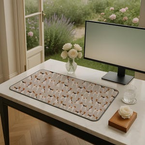 May include: A rectangular desk mat featuring a repeating pattern of terrier dogs in black, brown, and white on a light grey background. The mat has a black border and is on a white desk. A computer monitor, flowers, and a glass of water are also present.