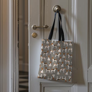 Puede incluir: Un bolso tote gris con un patrón repetido de perros marrones, negros y blancos. El bolso tiene asas negras y está colgado de una puerta. Los perros están en varias poses, creando un diseño lúdico.