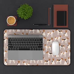 May include: A desk setup featuring a laptop, mouse, and a mousepad with a repeating pattern of Fox Terrier dogs in brown, black, and white. A cup of coffee, a small plant, and stationery are also on the dark surface.