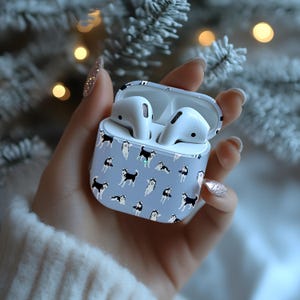Könnte beinhalten: Hellblaues AirPods-Case mit einem Muster aus schwarz-weißen Husky-Hunden. Das Case ist geöffnet und zeigt zwei weiße AirPods. Der Hintergrund ist verschwommen, mit einem weichen Fokus auf einem weißen Pullover und einem Weihnachtsbaum.
