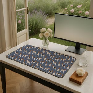 May include: A rectangular desk mat featuring a repeating pattern of dogs in brown, black, and white on a blue background. The mat is on a white desk with a computer monitor, a glass of water, and a vase of white roses.