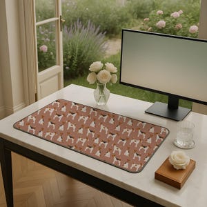May include: A rectangular desk mat featuring a repeating pattern of terrier dog illustrations in brown, black, and white on a dusty rose background. The mat is on a white desk with a computer monitor, flowers, and a glass of water.