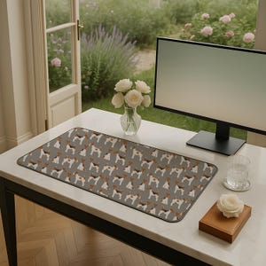 May include: A rectangular desk mat featuring a repeating pattern of dogs in brown, black, and white on a grey background. The mat is placed on a white desk, alongside a computer monitor and a vase of white roses.