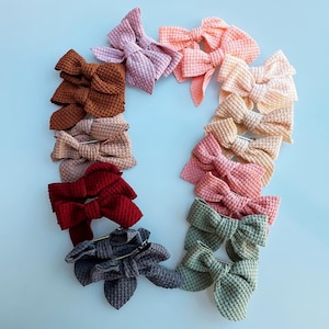May include: A collection of waffle-textured hair bows in various colors, including brown, pink, red, and green, arranged on a light blue surface. Each bow is secured with a metal clip, offering a range of styling options.