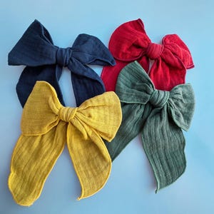 Puede incluir: Un surtido de lazos grandes de tela en varios colores, incluyendo azul marino, rojo, verde oliva y amarillo mostaza. Los lazos están dispuestos sobre una superficie azul claro, mostrando su apariencia texturizada y diseño sencillo. Accesorios para el cabello.