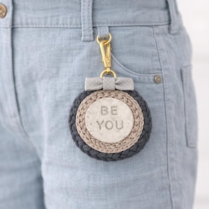 Puede incluir: Un llavero redondo con un diseño trenzado gris, beige y gris oscuro. El centro del llavero tiene las palabras "BE YOU" impresas. Un cierre dorado y un lazo gris con lunares blancos están adjuntos.