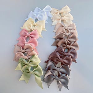 Op de afbeelding: Een verzameling kleurrijke haarstrikken, gerangschikt in een halve cirkel. De strikken zijn in verschillende pastelkleuren, waaronder wit, geel, roze, groen en bruin. Elke strik is gemaakt van een geribbelde stof en is in een klassieke strikvorm geknoopt.