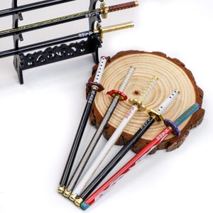 May include: Miniature decorative katana swords in various colors, including black, red, and white, with gold-colored accents. The swords are displayed on a wooden stand and a wooden slice. Each sword has Japanese text on the blade.