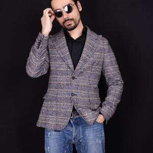 May include: A plaid blazer in shades of gray, blue, and brown, paired with a black shirt and blue jeans. The blazer has a classic cut with notched lapels and buttoned cuffs. The person is wearing sunglasses.