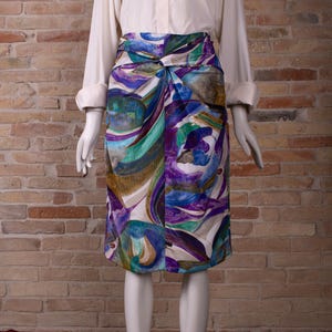 Op de afbeelding: Een kleurrijke rok tot op de knie met een uniek knoopdetail in de taille. De rok heeft een levendig abstract patroon in tinten blauw, paars, groen en wit. De rok wordt gecombineerd met een witte blouse met lange mouwen. De rok is ongeveer 60 cm lang.