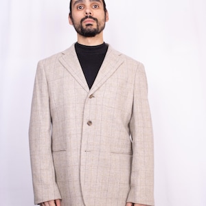 Könnte beinhalten: Beiger Blazer mit dezentem Karomuster. Der Blazer hat ein fallendes Revers, zwei Vordertaschen und einen Zwei-Knopf-Verschluss. Die Person im Bild trägt einen schwarzen Rollkragenpullover. Ein Blazer im klassischen Stil.