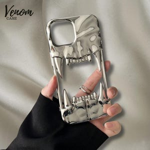 Peut inclure: Coque de téléphone argentée avec un motif de crocs. Le boîtier a une finition brillante et comporte le texte "Venom CASE" dans le coin supérieur gauche. Le design comprend de grandes canines et des dents pointues.