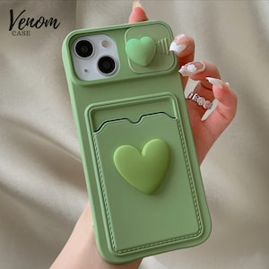 May include: A pale green phone case with a heart motif. The case has a cardholder and a camera cover, each with a heart. The text "Venom CASE" is printed in the upper left corner.