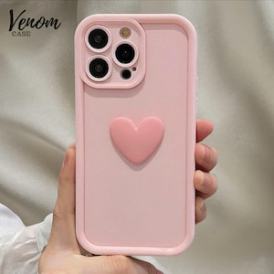 May include: A light pink phone case with a raised heart design. The case has a square camera cutout and the text "Venom Case" in the upper left corner. The case is held in a hand.