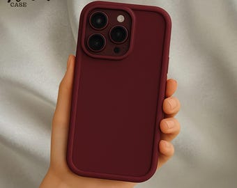 May include: A burgundy phone case with a smooth finish, held in a hand. The case has a raised border around the camera lenses and a matching frame. The text "Venom CASE" is in the upper left corner.