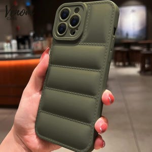 Pode incluir: Capa de telefone verde-oliva com design acolchoado. A capa tem um protetor de lente de c&acirc;mera e o texto "Venom Case". A capa &eacute; segurada em uma m&atilde;o.