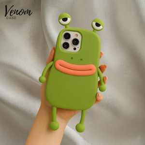May include: Green phone case with a cartoon monster design. The case has a green body, orange mouth, and two googly eyes. The case has arms and legs. The text "Venom Case" is in the upper left corner.
