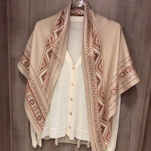 Op de afbeelding: Een beige sjaal met een geometrisch patroon in bruin-, wit- en donkerrode tinten, gedrapeerd over een crèmekleurig vest met knopen. De sjaal heeft een brede rand met gedetailleerde patronen, terwijl het vest een eenvoudige V-hals heeft.