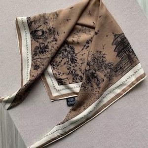 May include: A square scarf in a light brown shade featuring a dark brown printed design of a traditional pagoda, trees, and figures. The scarf has a cream-coloured border with a dark brown rope-like pattern and a black care label attached.