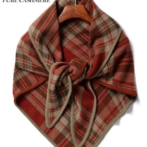 May include: A square scarf featuring a red, brown, and beige plaid pattern with a solid beige border. The scarf is tied in a knot and hangs from a wooden hanger. The text PURE CASHMERE and a ram head logo are visible in the upper left corner.