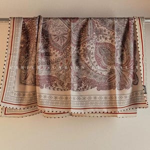 May include: A rectangular scarf with a paisley pattern in shades of burgundy, gray, and cream. The scarf has a cream border with a red stripe and a black dotted trim. The scarf is draped over a metal bar.