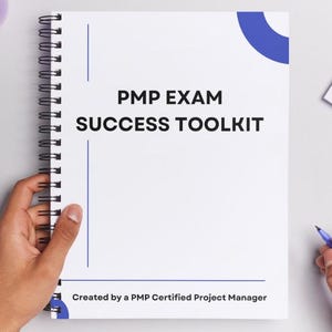 May include: A spiral-bound notebook with the text "PMP EXAM SUCCESS TOOLKIT" on the cover. A purple pen and a purple cup are on the white surface. A purple binder clip holds a small stack of white paper.