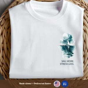 Puede incluir: Camiseta blanca con un diseño de acuarela de un velero en el mar y el texto "SAIL MORE. STRESS LESS." La camiseta está doblada sobre una superficie tejida.