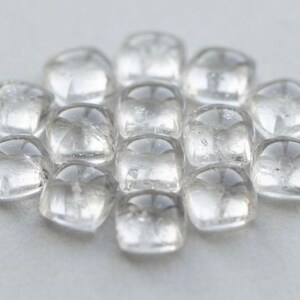 Puede incluir: Un conjunto de doce cabujones pequeños, transparentes y de forma cuadrada con bordes redondeados y una superficie lisa y pulida. Cada pieza es translúcida, refleja la luz y está dispuesta en un grupo circular compacto sobre una superficie neutra.