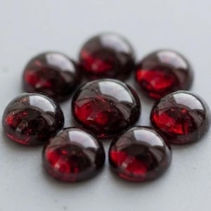 May include: Seven smooth, round, deep red cabochon gemstones arranged in a circular flower-like pattern. Each stone features a polished, domed surface that reflects light, showcasing a consistent dark crimson hue and a glossy, reflective finish.
