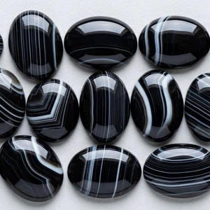 Op de afbeelding: Een verzameling van vijftien gepolijste, ovaalvormige zwarte onyx edelstenen met opvallende, dunne witte bandpatronen. Elke steen heeft een glad, reflecterend oppervlak met unieke lineaire en gebogen witte strepen over de zwarte basis.