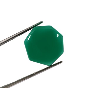 May include: A polished, hexagonal emerald green gemstone, held by silver tweezers against a white backdrop. The gem's facets catch the light, highlighting its smooth surface. This gemstone is suitable for jewelry making and is approximately 2.5 cm across.