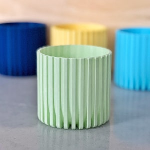 Puede incluir: Cuatro macetas cilíndricas acanaladas de colores vivos. La maceta en primer plano es verde claro, la maceta de la derecha es azul claro, la maceta de la izquierda es azul oscuro y la maceta del fondo es amarilla.