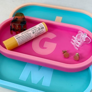 May include: Two oval-shaped trays, one pink and one blue, with raised letters "G" and "M" on the surface. The pink tray contains a yellow lip balm tube, a brown hair clip, two gold earrings, and a clear plastic elephant figurine. The blue tray is stacked on top of the pink tray.