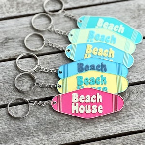 Puede incluir: Varios llaveros coloridos con las palabras "Beach" o "Beach House" en letras blancas. Los llaveros son de color azul, amarillo y rosa, con anillas y cadenas plateadas. Están colocados sobre una superficie de madera envejecida.