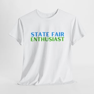 May include: A white t-shirt featuring the words "STATE FAIR ENTHUSIAST" in blue and green block letters. The shirt has a crew neck and is made of a soft material. This casual t-shirt is suitable for everyday wear.