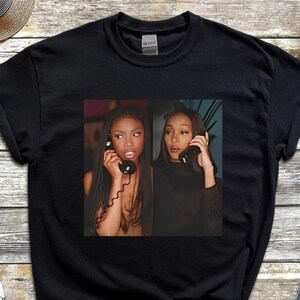 May include: Black t-shirt featuring a graphic of two women on the phone. The image is in a rectangular frame. The shirt is displayed on a wooden surface with a pair of black and white sneakers, a straw hat, and denim shorts.