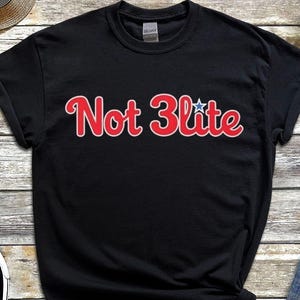 May include: Black crew neck t-shirt with the text "Not 3lite" in red with a white outline and a blue star. The shirt is paired with denim shorts, black sneakers, a straw hat, and a smartwatch.