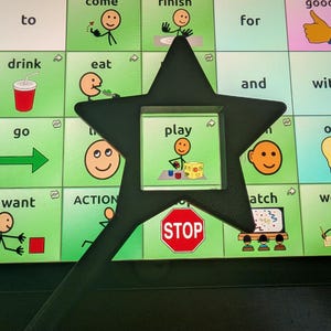 May include: A black star-shaped magnifying tool positioned over a communication board featuring various icons and text labels including drink, eat, play, and stop. The tool highlights the central play icon showing a stick figure with blocks.