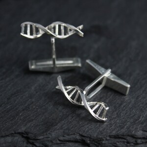 Pu&ograve; includere: Un paio di gemelli d'argento con un design a doppia elica di DNA 3D. I gemelli sono su una superficie grigio scuro.