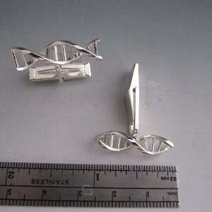 Pu&ograve; includere: Un paio di gemelli d'argento con un design a doppia elica di DNA. I gemelli sono mostrati accanto a un righello per riferimento alle dimensioni.