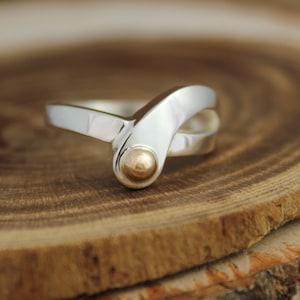 May include: A silver ring with a gold accent in the shape of a sphere. The ring is designed with a curved, overlapping band.