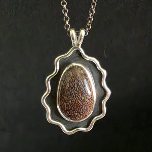 May include: A silver pendant necklace featuring a large, oval-shaped stone with a brown and tan patterned surface. The stone is set within a silver frame with a wavy edge, suspended from a silver chain.