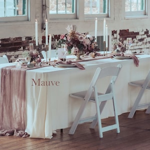 May include: A white table with a white tablecloth and a mauve runner. The table is set with candles, plates, and glasses. There are white folding chairs on either side of the table. The table is in a room with white brick walls and large windows.