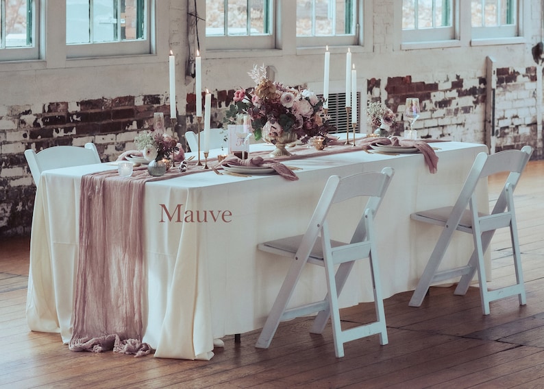 May include: A white table with a white tablecloth and a mauve runner. The table is set with candles, plates, and glasses. There are white folding chairs on either side of the table. The table is in a room with white brick walls and large windows.
