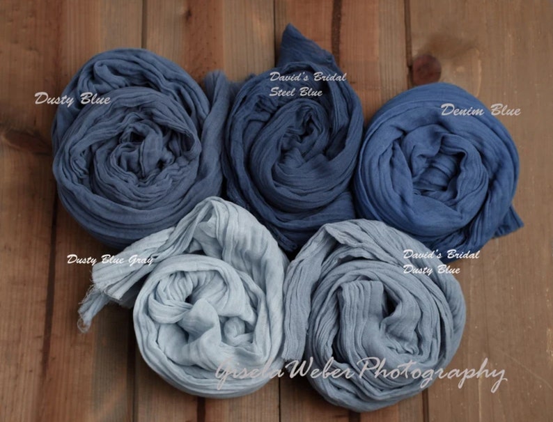 May include: Four rolls of blue fabric in different shades of blue, including dusty blue, steel blue, denim blue, and dusty blue gray. The fabric is arranged in a circle on a wooden surface. The text "David's Bridal" and "Gisela Weber Photography" is visible in the image.