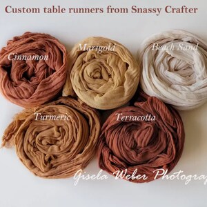 May include: Six rolled fabric table runners in various shades of brown, including cinnamon, marigold, beach sand, turmeric, terracotta, and a lighter brown. The fabric is a soft, gauzy material. The text "Custom table runners from Snassy Crafter" and "Gisela Weber Photography" is visible in the image.