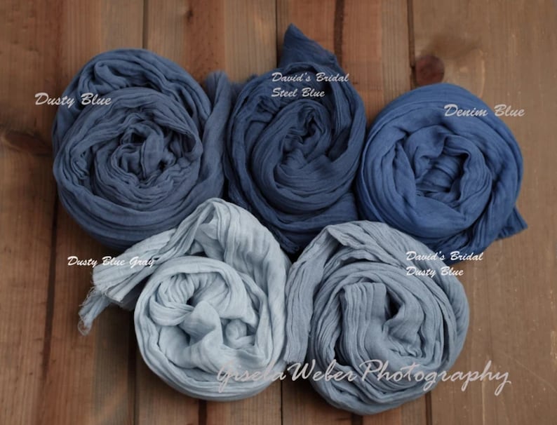May include: Four rolls of blue fabric in different shades of blue, including dusty blue, steel blue, denim blue, and dusty blue gray. The fabric is arranged in a circle on a wooden surface. The text "David's Bridal" and "Gisela Weber Photography" is visible in the image.
