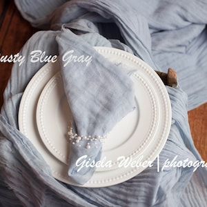 May include: A dusty blue gray fabric napkin is folded and placed on a white plate with a beaded napkin ring. The plate is on top of a larger white plate. The fabric is draped over a wooden surface.