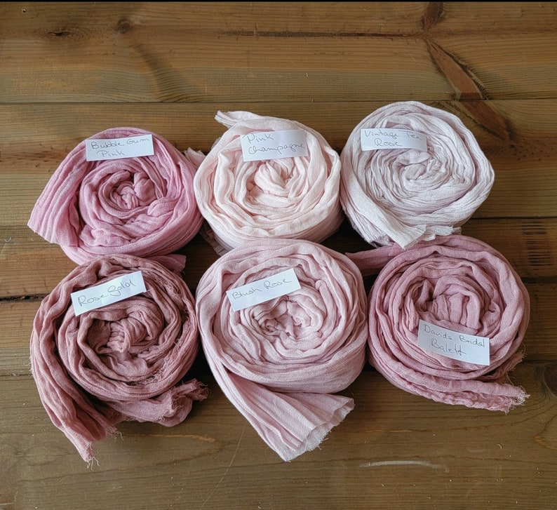May include: Six spools of pink fabric dyed in different shades of pink. The fabric is rolled up and labeled with the names of the colors: "Bubble Gum Pink", "Pink Champagne", "Vintage Tea Rose", "Rose Petal", "Blush Rose", and "David's Bridal Ballet".